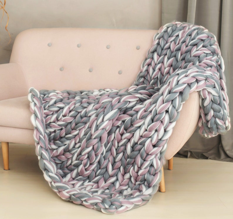 K&ouml;nnte beinhalten: Eine dicke Strickdecke in den Farben Grau, Wei&szlig; und Rosa, &uuml;ber ein hellrosa Sofa gelegt.
