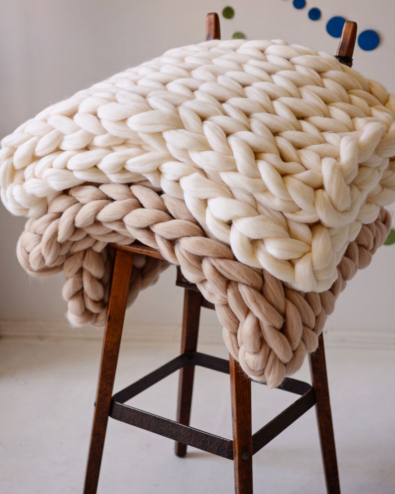 May include: A chunky knit throw blanket in two shades of beige, draped over a wooden stool. The blanket is made of thick yarn and has a soft, cozy texture.