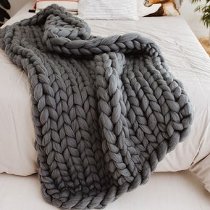 May include: A chunky knit gray blanket draped over a bed. The blanket's thick, woven texture is prominently displayed. Pillows with various patterns and colors are visible in the background, adding to the cozy aesthetic. The bed is covered with a neutral-toned sheet.