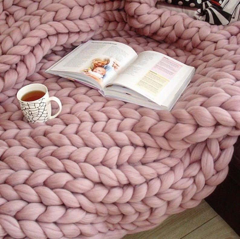 Op de afbeelding: Een zachte, dikke gebreide deken in een stoffige roze kleur. De deken is gemaakt van dik garen en heeft een kabelbreipatroon. Een kopje thee en een boek liggen op de deken.