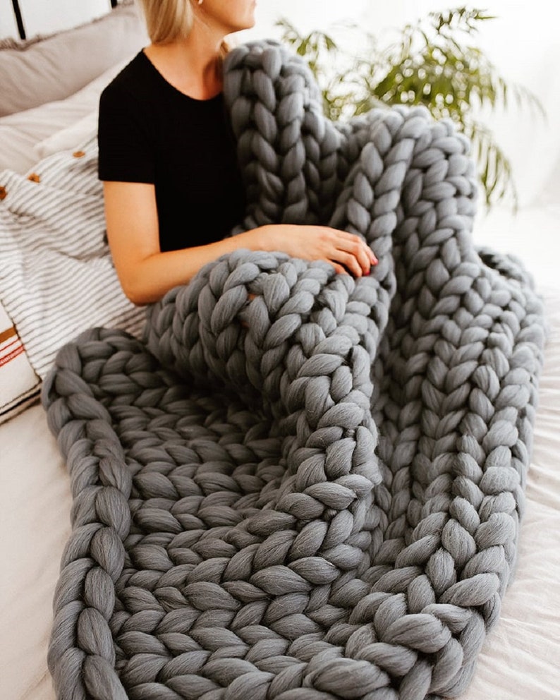 K&ouml;nnte beinhalten: Eine gro&szlig;e, grob gestrickte graue Wolldecke aus dickem Garn. Die Decke liegt &uuml;ber einem Bett und eine Person sitzt auf dem Bett.