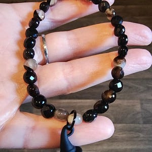 May include: A beaded bracelet featuring black and brown faceted beads with a silver accent bead. A black witch hat charm dangles from the bracelet. The bracelet is shown on a hand.