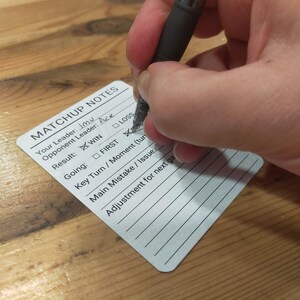 May include: A white Matchup Notes card with the text "MATCHUP NOTES" and lines for notes. A hand is holding a black pen and marking the "WIN" box. The card is on a wooden surface.