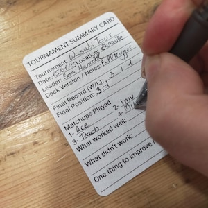 May include: A white tournament summary card with black text is being filled out with a pen. The card includes fields for tournament details, final record, and matchups. The text on the card reads "Wasabi Tour" and "Fulltrigger".