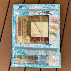 Pode incluir: Uma bolsa de projeto de tecido azul com uma janela de plástico transparente que mostra um bloco de colcha de retalhos. A bolsa tem um fecho de correr e uma etiqueta que diz "Bloco 6". Também há vários carretéis de fio de bordar e um pacote de agulhas dentro da bolsa.