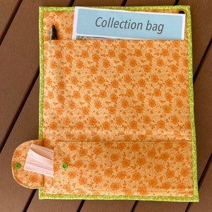 May include: A brown fabric collection bag with a green trim and a floral pattern. The bag has a pocket on the front with the text "Collection bag" printed on a light blue piece of paper. The bag also has a smaller pocket on the bottom with a white fabric insert.