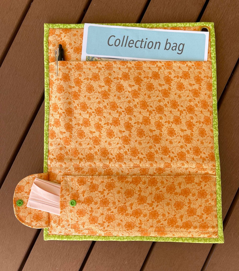 May include: A brown fabric collection bag with a green trim and a floral pattern. The bag has a pocket on the front with the text "Collection bag" printed on a light blue piece of paper. The bag also has a smaller pocket on the bottom with a white fabric insert.