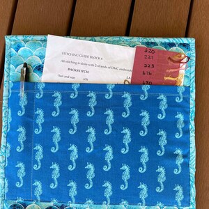 May include: A blue fabric project with a pocket containing a pen and a piece of paper with instructions for stitching guide block 6. The fabric has a pattern of white seahorses on a blue background. The paper has the text "STITCHING GUIDE BLOCK 6", "All stitching is done with 2 strands of DMC embroidery", "BACKSTITCH", "Sun and star 676", "420", "221", "223", "676", and "670".
