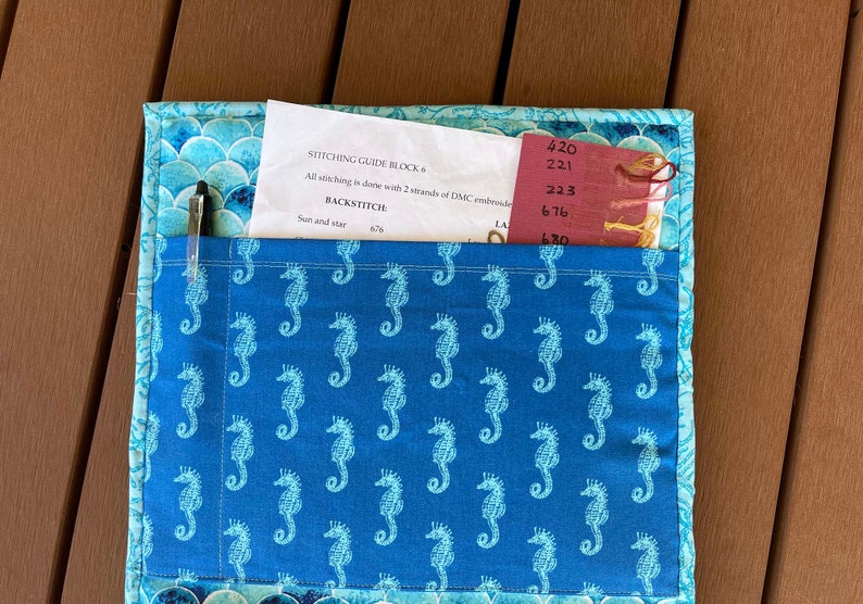 May include: A blue fabric project with a pocket containing a pen and a piece of paper with instructions for stitching guide block 6. The fabric has a pattern of white seahorses on a blue background. The paper has the text "STITCHING GUIDE BLOCK 6", "All stitching is done with 2 strands of DMC embroidery", "BACKSTITCH", "Sun and star 676", "420", "221", "223", "676", and "670".