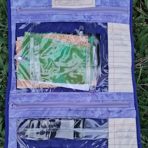 May include: A purple quilted bag with clear vinyl pockets. The pockets contain fabric scraps and a pen. The bag has a label that says "Chloe's Bag". The pockets are labeled "Vinyl lining Hardware" and "Black Strap Hardware".