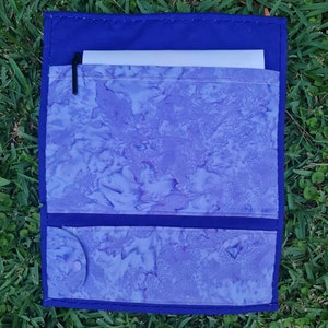 May include: A blue fabric pouch with a white and purple patterned lining. The pouch has a flap with a pocket for a pen or pencil. The pouch is closed with a button.