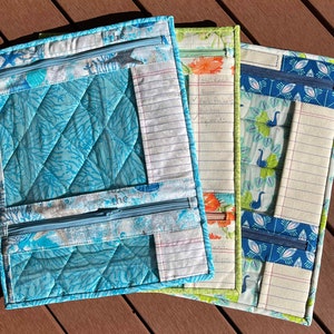 May include: Three zippered pouches made of fabric with different patterns. The pouches have a clear plastic window and a zippered pocket. The pouches are lined with white paper with blue lines. The text "the" is written on the front of the pouches. The text "still need" is written on the middle pouch with a list of items.