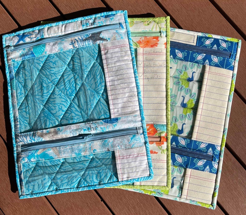 May include: Three zippered pouches made of fabric with different patterns. The pouches have a clear plastic window and a zippered pocket. The pouches are lined with white paper with blue lines. The text "the" is written on the front of the pouches. The text "still need" is written on the middle pouch with a list of items.