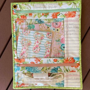 May include: A green and floral fabric zippered pouch with clear plastic pockets. The pouch is labeled "Travel Collection Bag" and has a handwritten list of items needed for the project, including "foam", "back pockets", "binding", and "zip ends".