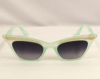 Könnte beinhalten: Mintgrüne Cat-Eye-Sonnenbrille mit goldenen Kettendetails am oberen Rand des Rahmens. Die Gläser haben einen Farbverlauf von Dunkelgrau zu Hellgrau. Die Bügel sind in passendem Mintgrün gehalten.