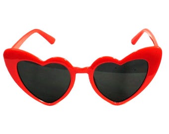 May include: Bright red sunglasses featuring heart-shaped frames and dark tinted lenses. The plastic frames are molded into a distinct heart shape with matching red temples extending from the sides, creating a bold and uniform appearance.
