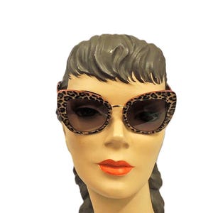 May include: Cat-eye sunglasses with a leopard print pattern on the frames and temples. The lenses are a gradient brown color. The sunglasses are displayed on a mannequin head with brown hair and orange lipstick.