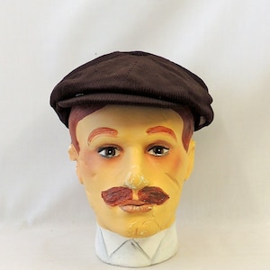 Puede incluir: Una gorra de estilo newsboy de pana marrón descansa sobre una cabeza de maniquí pintada con bigote y pelo rojo. El maniquí lleva una camisa blanca con cuello. El fondo es blanco.