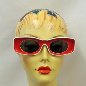 May include: A white mannequin head with a red and white pair of sunglasses. The mannequin is wearing a bright orange top with white flowers.