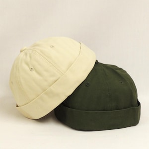 May include: Two cotton docker hats. One is a light beige colour, and the other is a dark olive green. The hats are folded at the brim and have a rounded crown. They are resting on a white background.