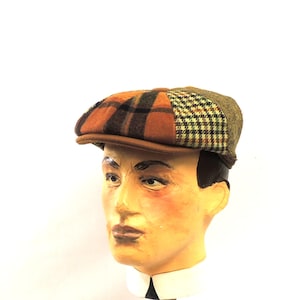 May include: A patchwork newsboy cap with a brown leather brim. The cap features a mix of plaid and tweed patterns in shades of brown, orange, and green. The hat is displayed on a mannequin head with a white collar.