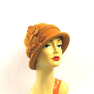 May include: Mustard-colored cloche hat with floral embellishments. The hat features a rounded crown and a small brim. The flowers are clustered on the side of the hat. The hat is displayed on a mannequin head with an orange and white floral top.