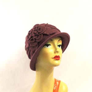 May include: A burgundy cloche hat with a floral design. The hat has a rounded crown and a small brim. It is displayed on a mannequin head wearing an orange top with white floral patterns. The hat is a deep, rich colour.