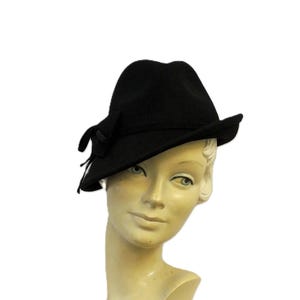 Peut inclure: Un chapeau fedora noir avec un petit nœud sur le côté. Le chapeau a un design classique avec un bord légèrement incurvé. Le chapeau est présenté sur une tête de mannequin au teint neutre, sur fond blanc.