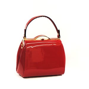 Puede incluir: Un bolso rojo brillante con una forma estructurada y un asa superior. El bolso presenta herrajes dorados y un cierre de broche. El asa es negra y el bolso está sobre un fondo blanco.