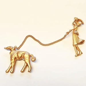 Könnte beinhalten: Goldfarbene Brosche mit einem stilisierten Hund an einer Kette, verbunden mit einer Figur einer Person mit Hut und Kleid. Der Hund ist eine schlanke Rasse, und die Person hält die Kette. Die Brosche hat eine Vintage-Ästhetik.