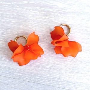 Könnte beinhalten: Ein Paar Creolen mit leuchtend orangefarbenen, geschichteten blütenblattartigen Akzenten. Die Ohrringe bestehen aus einer kreisförmigen, goldfarbenen Metall-Creole, an der die lebendigen, halbtransparenten orangefarbenen Blütenteile befestigt sind.