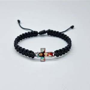 以下が含まれることがあります： イエスのカラフルな画像が描かれた銀色の十字架チャームが付いた、黒い編み込みブレスレットです。