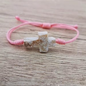 Op de afbeelding: Een roze koordarmband met een witte stenen kruisbedel. Het kruis heeft een ruwe, natuurlijke textuur.