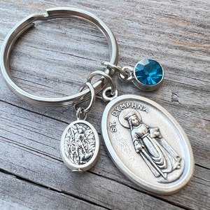 May include: Silver keychain with three charms. One charm is a small oval with a detailed image, another is an oval pendant with the text "ST. DYMPNA" and an image of a saint, and a blue crystal. The keychain is on a wooden surface.