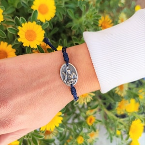 May include: A navy blue cord bracelet with a silver charm featuring a religious figure. The charm is oval-shaped and has a detailed design.