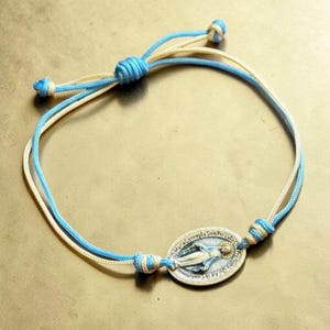 Könnte beinhalten: Verstellbares Armband mit einem silbernen, ovalen Anhänger mit einer religiösen Figur und Text. Das Armband hat eine blau-cremefarbene Kordel mit verstellbaren Knoten. Der Anhänger befindet sich in der Mitte der Kordel.