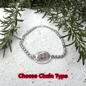 Puede incluir: Una pulsera de plata con un colgante ovalado detallado que presenta una escena religiosa y la palabra "Italy". La pulsera tiene un dise&ntilde;o de eslabones de cadena y se muestra sobre una superficie blanca con follaje verde. El texto "Choose Chain Type" est&aacute; en la parte inferior.