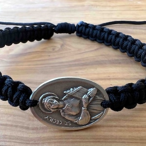 Op de afbeelding: Een armband van zwart gevlochten koord met een ovale zilveren bedel met een reliëfbeeld van een vrouw die een muziekinstrument vasthoudt. De bedel is gegraveerd met "ST CECILIA".