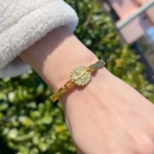 Könnte beinhalten: Ein goldfarbenes Armband mit einem runden Anhänger, der eine Taube zeigt. Das Armband befindet sich an einem Handgelenk.