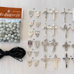 May include: A set of rosary beads with white and gray beads, black elastic cord, and a variety of silver metal charms. The charms include crosses, medallions, and images of religious figures. The charms are labeled with letters and numbers.