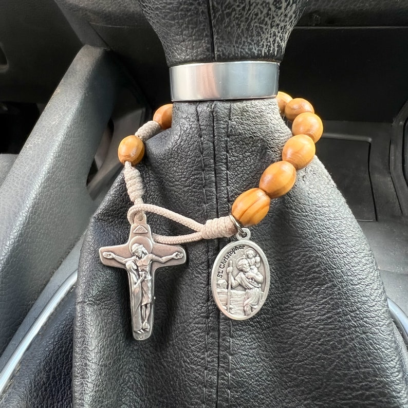 K&ouml;nnte beinhalten: Ein Rosenkranz aus Holz mit einem silbernen Kreuz und einem silbernen Anh&auml;nger mit einem Heiligen. Der Rosenkranz h&auml;ngt am Schalthebel eines Autos.