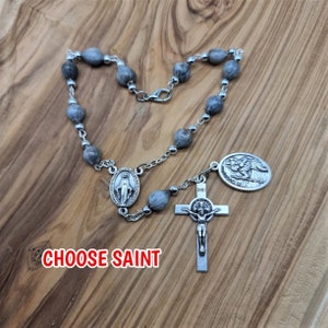 May include: A silver rosary with grey wooden beads, a silver crucifix, and a silver medal of Saint Jude Thaddeus. The text "CHOOSE SAINT" is visible below the rosary.