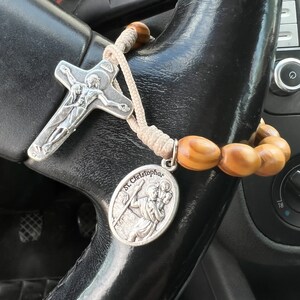 K&ouml;nnte beinhalten: Ein silbernes Rosenkranz mit einem Holzperlen-Anh&auml;nger und einem silbernen Anh&auml;nger mit einer Darstellung des Heiligen Christophorus. Der Rosenkranz h&auml;ngt am Lenkrad eines Autos.