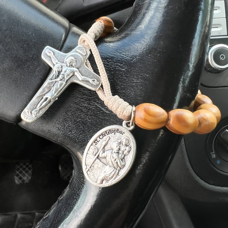 K&ouml;nnte beinhalten: Ein silbernes Rosenkranz mit einem Holzperlen-Anh&auml;nger und einem silbernen Anh&auml;nger mit einer Darstellung des Heiligen Christophorus. Der Rosenkranz h&auml;ngt am Lenkrad eines Autos.
