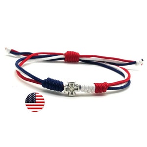 May include: A red, white, and blue braided bracelet with a silver cross charm. The bracelet is tied with a knot and has a sliding closure.
