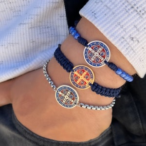Puede incluir: Tres pulseras, una con cadena de plata y dos con cordones azules y rojos, cada una con un dije redondo de plata con una cruz y la inscripción "S.M.Q.L.I.V.B.N.S.D.N.J.M.J.C.S.P.B.S.M.Q.L.I.V.B.N.S.D.N.J.M.J.C.S.P.B."
