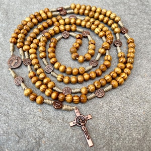 Può includere: Un rosario di legno con perline marroni e un crocifisso in rame. Le perline sono infilate su un cordoncino beige. Il crocifisso ha una piccola figura di Gesù.