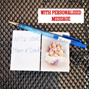 May include: A white card with the handwritten message "With love, Mam & Dad" next to a card with a picture of three angels. The text "With Personalized Message" is written in red on a white background.