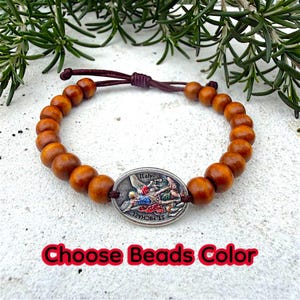 Puede incluir: Una pulsera de cuentas de madera con un dije ovalado plateado que representa una escena religiosa y la palabra "Italy". La pulsera tiene un cord&oacute;n marr&oacute;n y el texto "Choose Beads Color" en rojo.