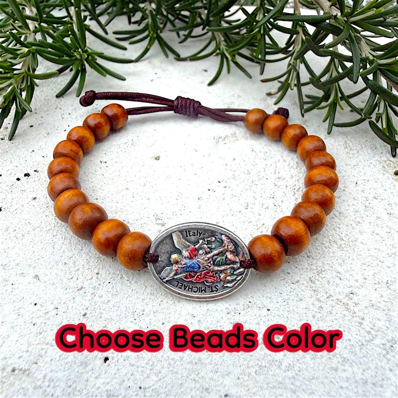 Puede incluir: Una pulsera de cuentas de madera con un dije ovalado plateado que representa una escena religiosa y la palabra "Italy". La pulsera tiene un cord&oacute;n marr&oacute;n y el texto "Choose Beads Color" en rojo.